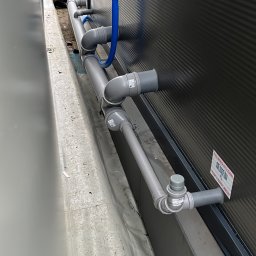 MGO Mateusz Gosz - Szare rury kanalizacyjne z niebieskim elastycznym przewodem przymocowane do ciemnej, żebrowanej ściany budynku, widoczne z boku, z elementami instalacji zewnętrznej w tle.