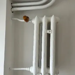 Biały, żeliwny grzejnik centralnego ogrzewania z rurami, zamontowany na ścianie. Widoczne ślady po malowaniu i niedokładności w wykończeniu.
