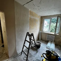Wnętrze pokoju w trakcie remontu: ściany pokryte siatką wzmacniającą pod tynk, drabina, wiadro z narzędziami, odkurzacz przemysłowy, widoczne przewody elektryczne w suficie, okno z widokiem na zieleń.