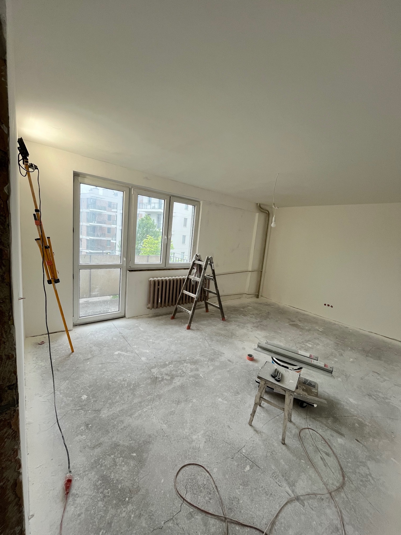 Pokój w trakcie remontu w Gdańsku: drabina, lampa budowlana i narzędzia na podłodze. Widoczne gładkie, białe ściany i okno balkonowe.