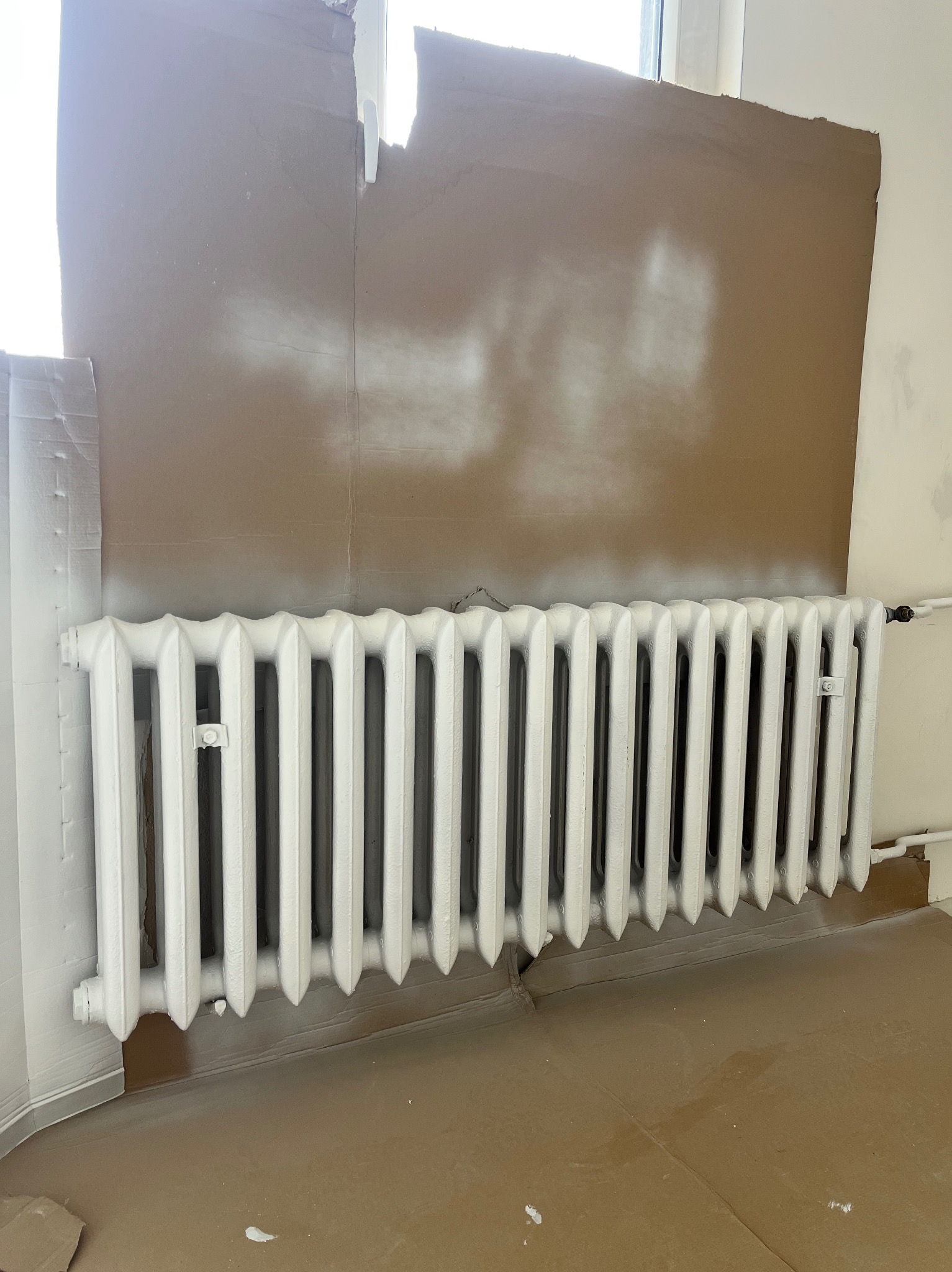 Biały, żeliwny grzejnik po malowaniu, zabezpieczony kartonem na ścianie i podłodze. Widoczne świeże ślady farby na ścianie. Oświetlenie dzienne.