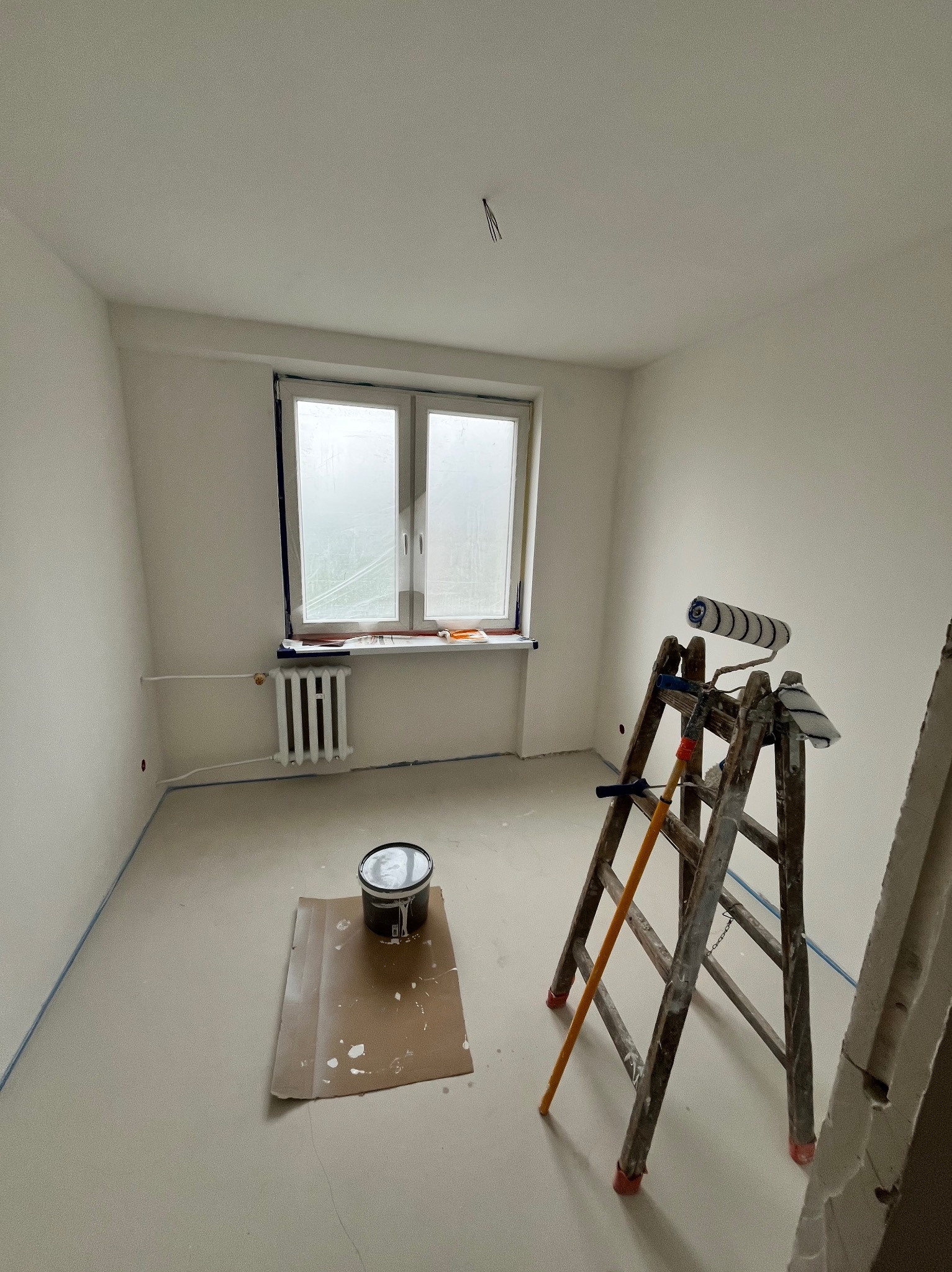 Pokój w trakcie malowania: drabina z wałkiem, puszka farby na kartonie, okno zabezpieczone folią, świeżo pomalowane ściany z niebieską taśmą malarską.