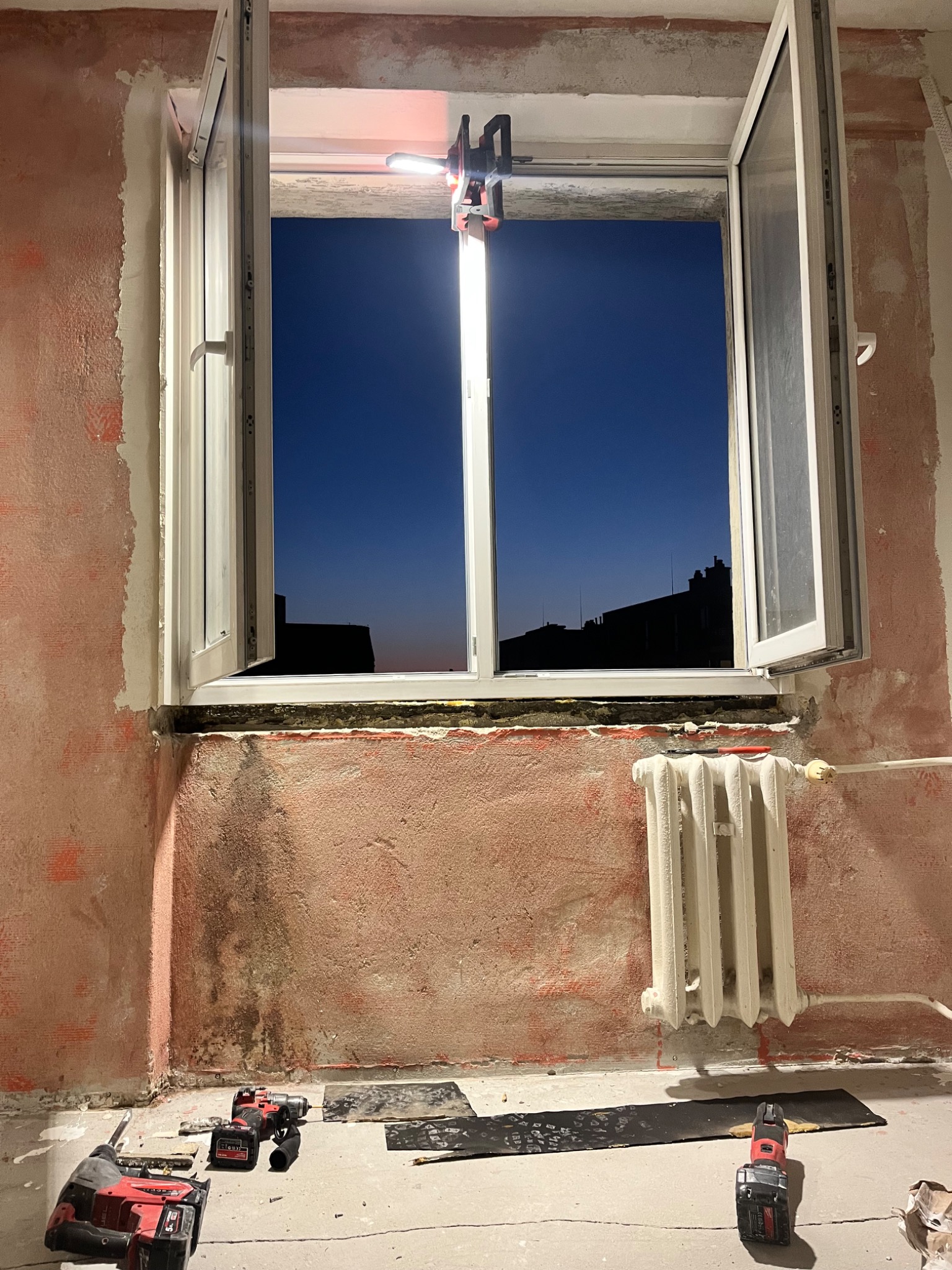 Montaż okna PCV z oświetleniem roboczym w surowym wnętrzu. Widoczne narzędzia i grzejnik. Ściany w trakcie przygotowania do wykończenia.
