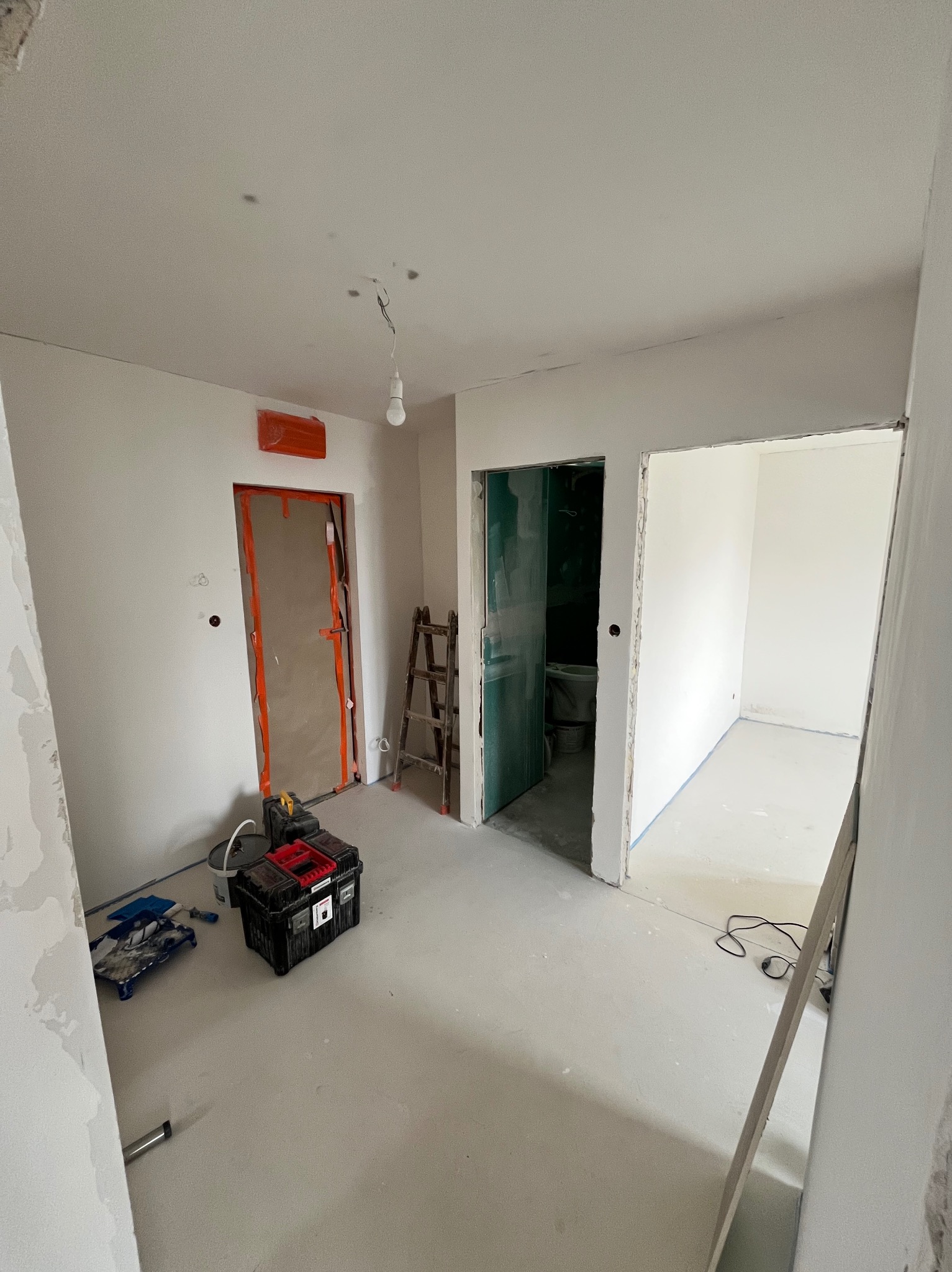Wnętrze w trakcie remontu: zabezpieczone drzwi, drabina, narzędzia malarskie, ściany przygotowane do malowania, surowe wykończenie, widok na łazienkę i kolejne pomieszczenie.