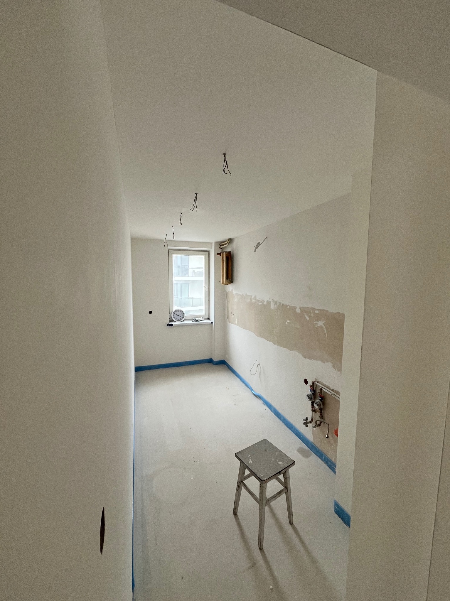 Wnętrze w trakcie remontu: białe ściany po szpachlowaniu, zabezpieczone podłogi i okna. Stary stołek na środku, widoczne kable i instalacja wodna.