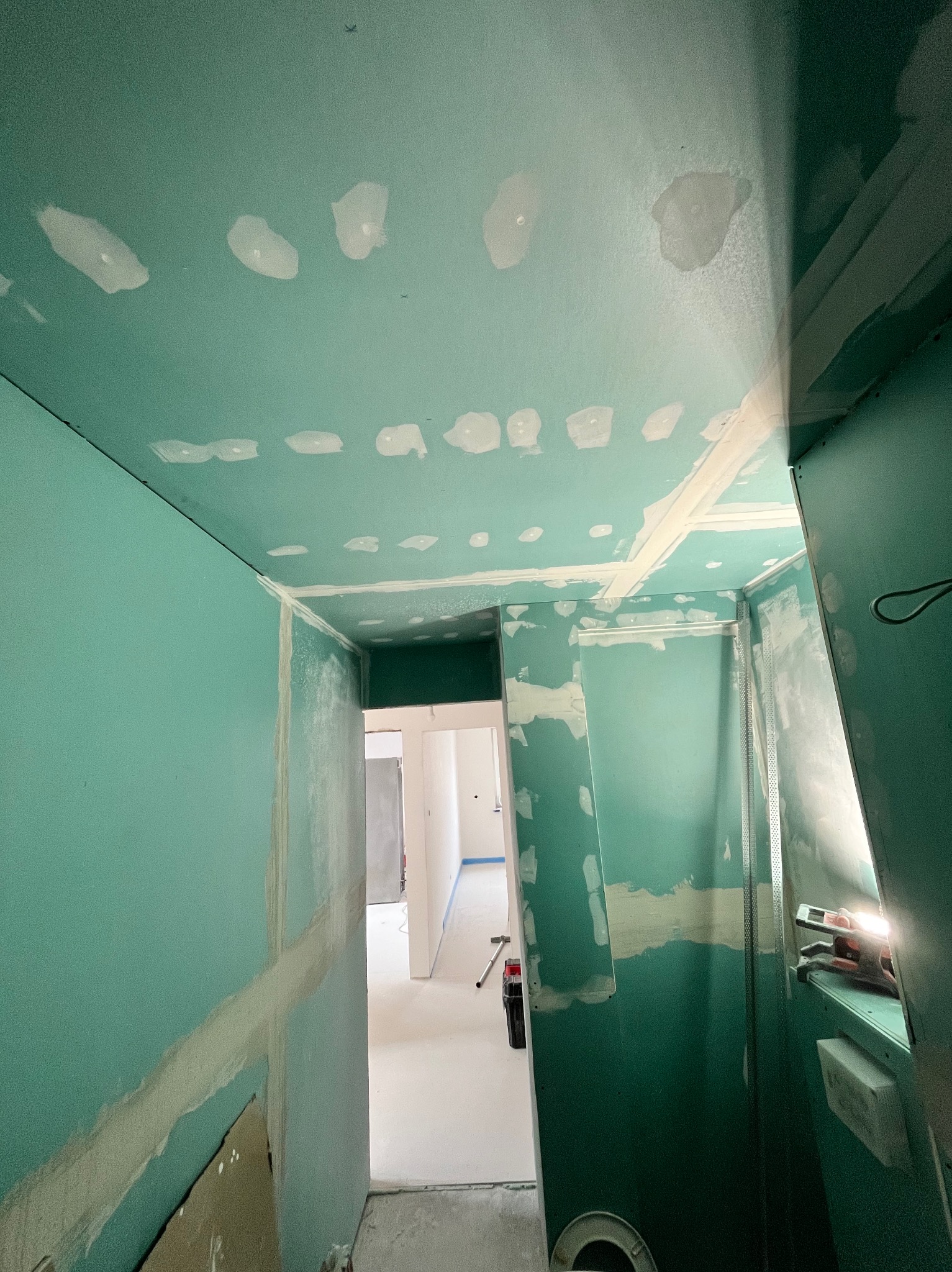 Zielone płyty karton-gips na ścianach i suficie, wstępnie wyszpachlowane, widoczne łączenia i wkręty. W tle otwarte drzwi do kolejnych pomieszczeń.