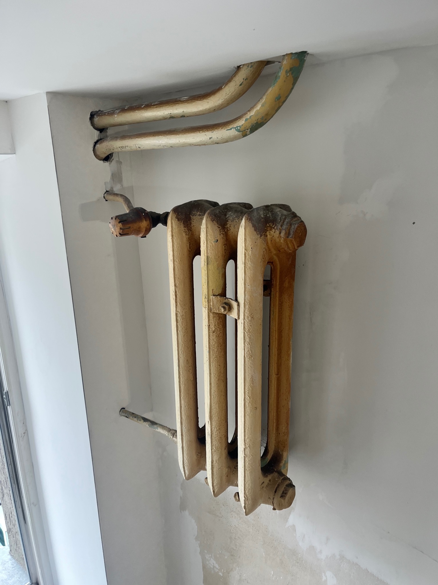 Żeliwny, beżowy grzejnik na tle białej ściany w trakcie remontu. Widoczne rury grzewcze i ślady szpachlowania. Ujęcie z dołu, ukazujące stan przed wykończeniem.