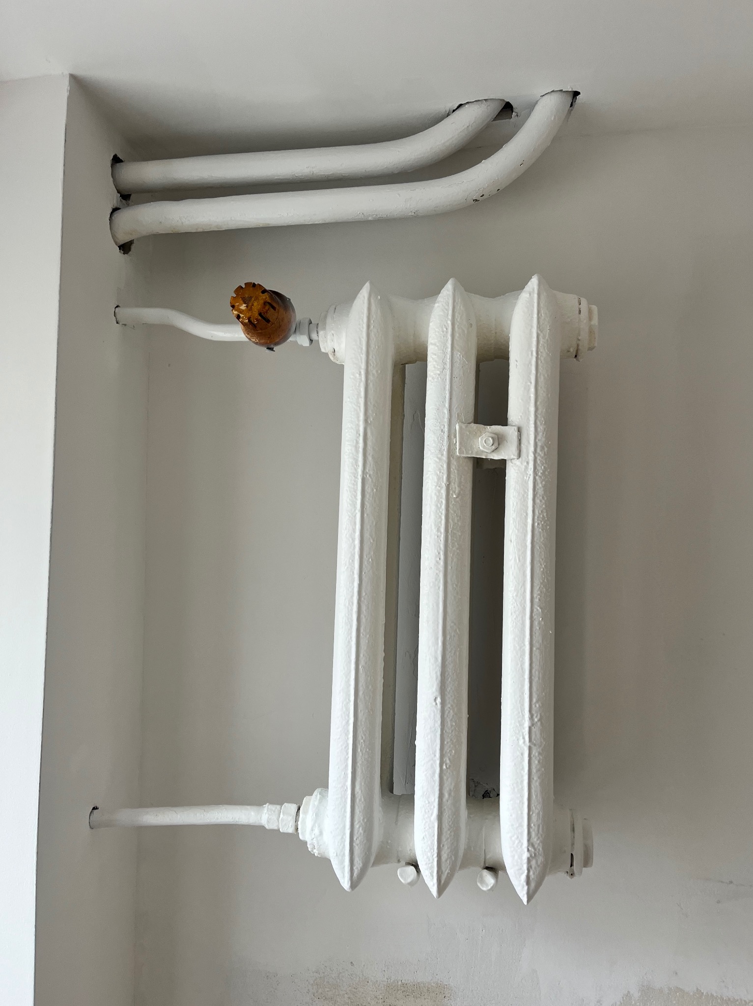 Biały, żeliwny grzejnik centralnego ogrzewania z rurami, zamontowany na ścianie. Widoczne ślady po malowaniu i niedokładności w wykończeniu.