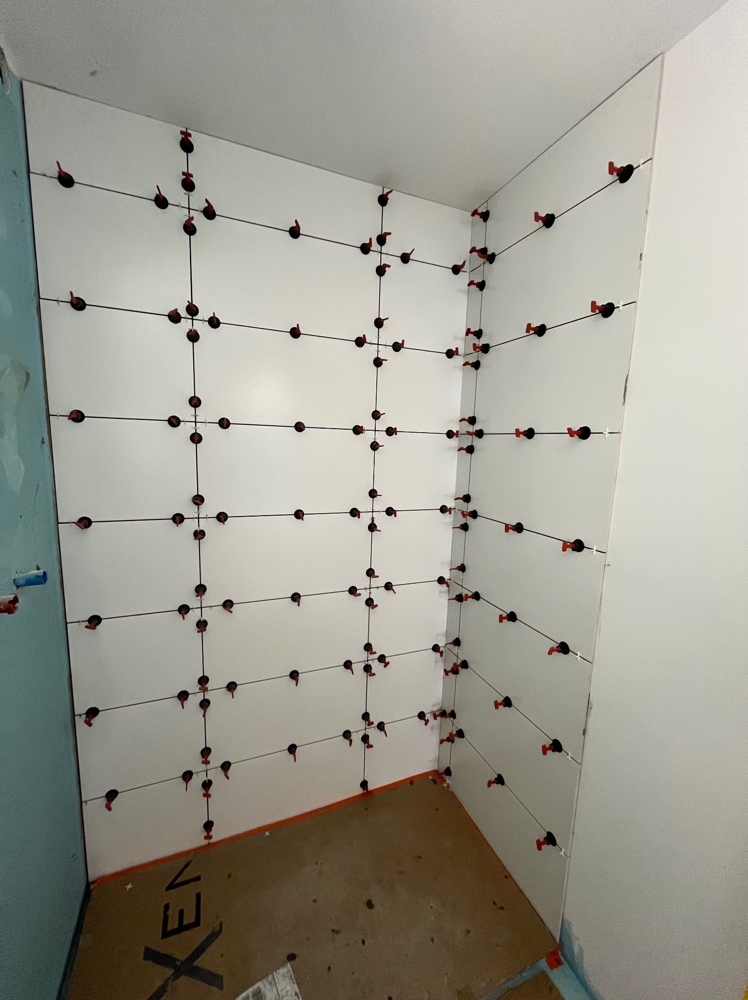 Białe płytki ścienne w trakcie układania, z użyciem poziomowania krzyżykami i klinami w łazience. Widoczny fragment podłogi i ściany w kolorze delikatnego błękitu.