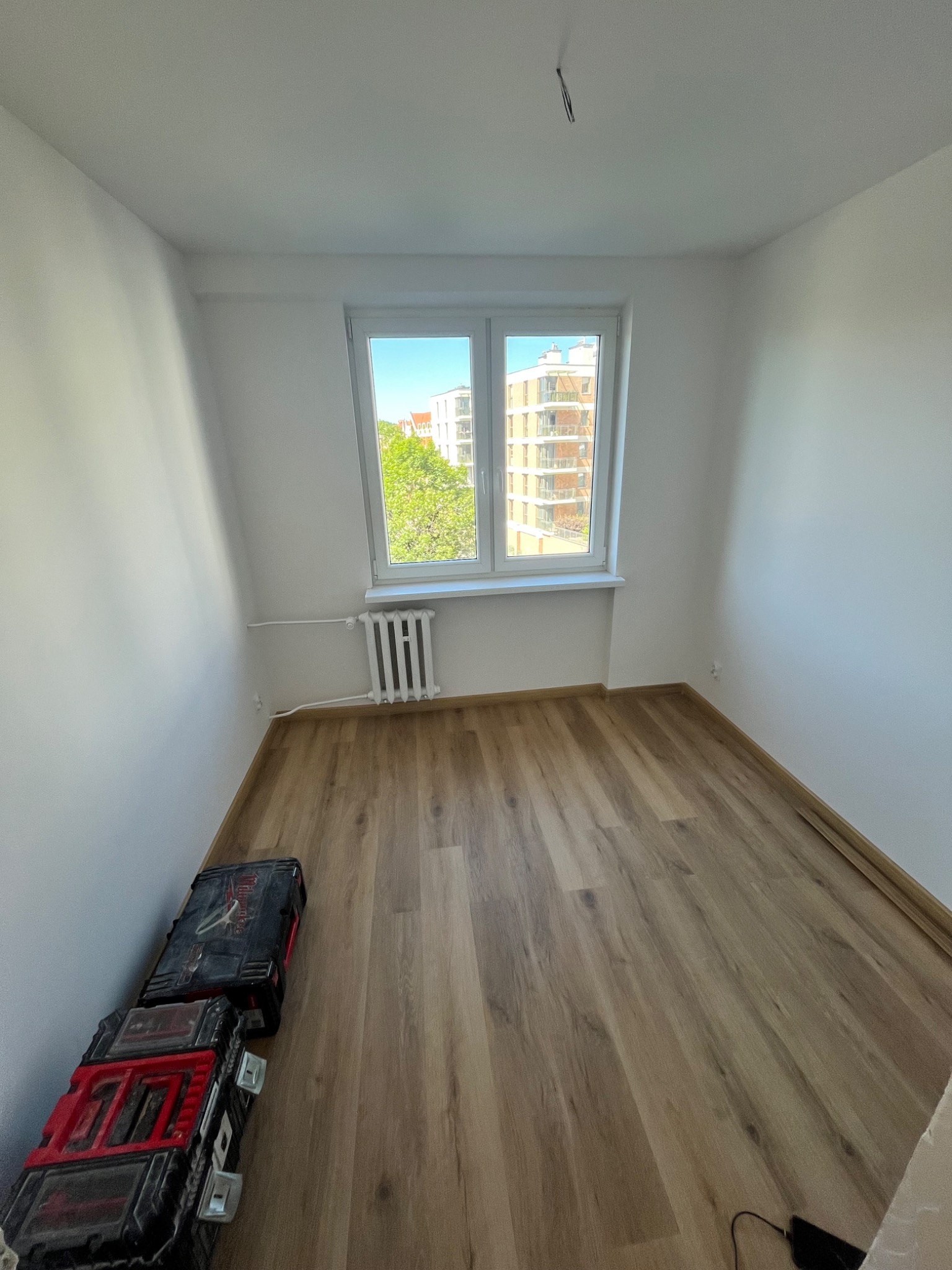 Pusty pokój po remoncie z panelami podłogowymi, białymi ścianami i oknem z widokiem na budynek. Skrzynki narzędziowe przy ścianie. Wiszące kable na suficie.