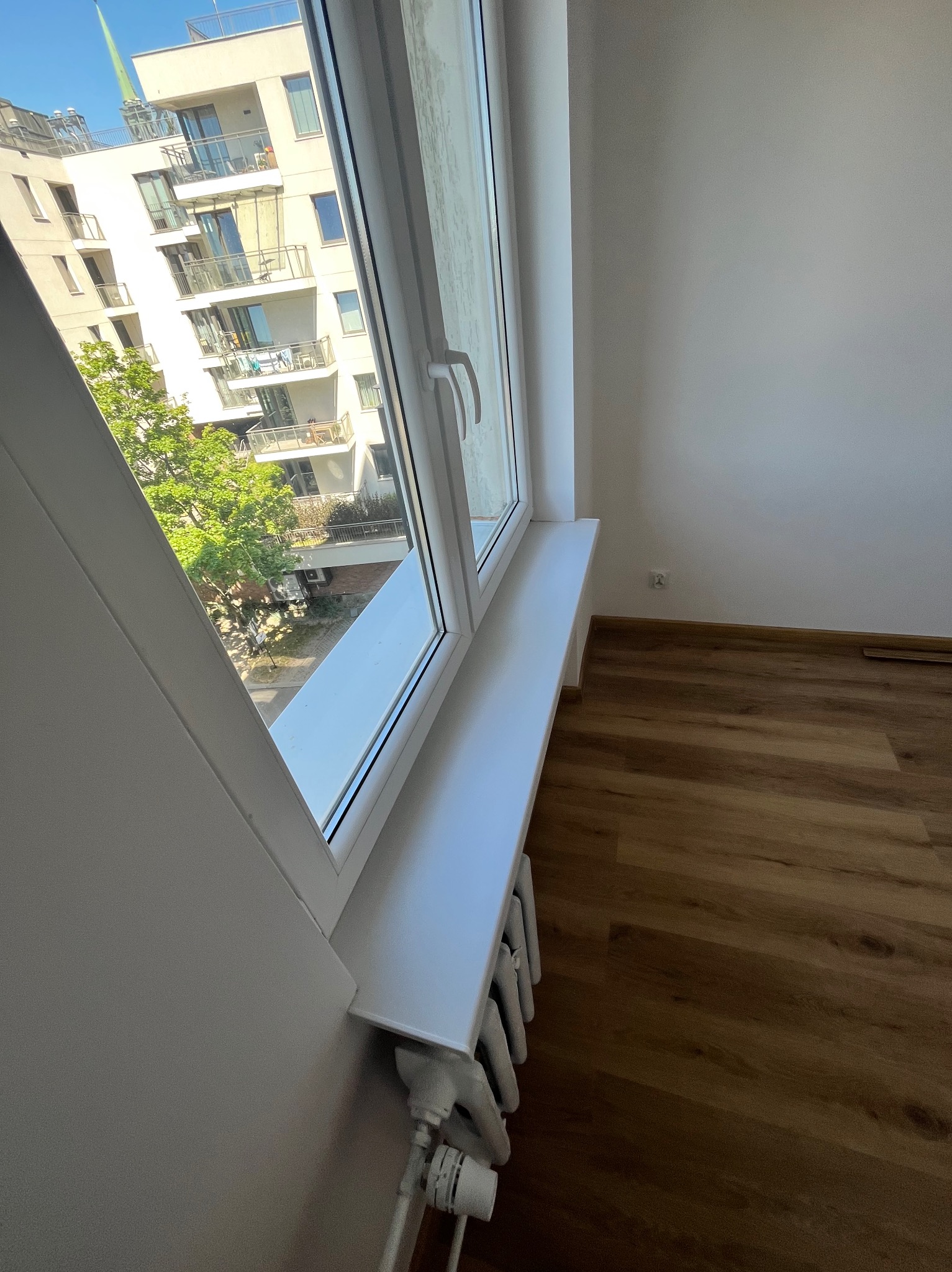 Białe okno PCV z widokiem na budynek mieszkalny i drzewa, zamontowane w pokoju z drewnianą podłogą i grzejnikiem. Świeżo wykończone ściany pomalowane na biało.