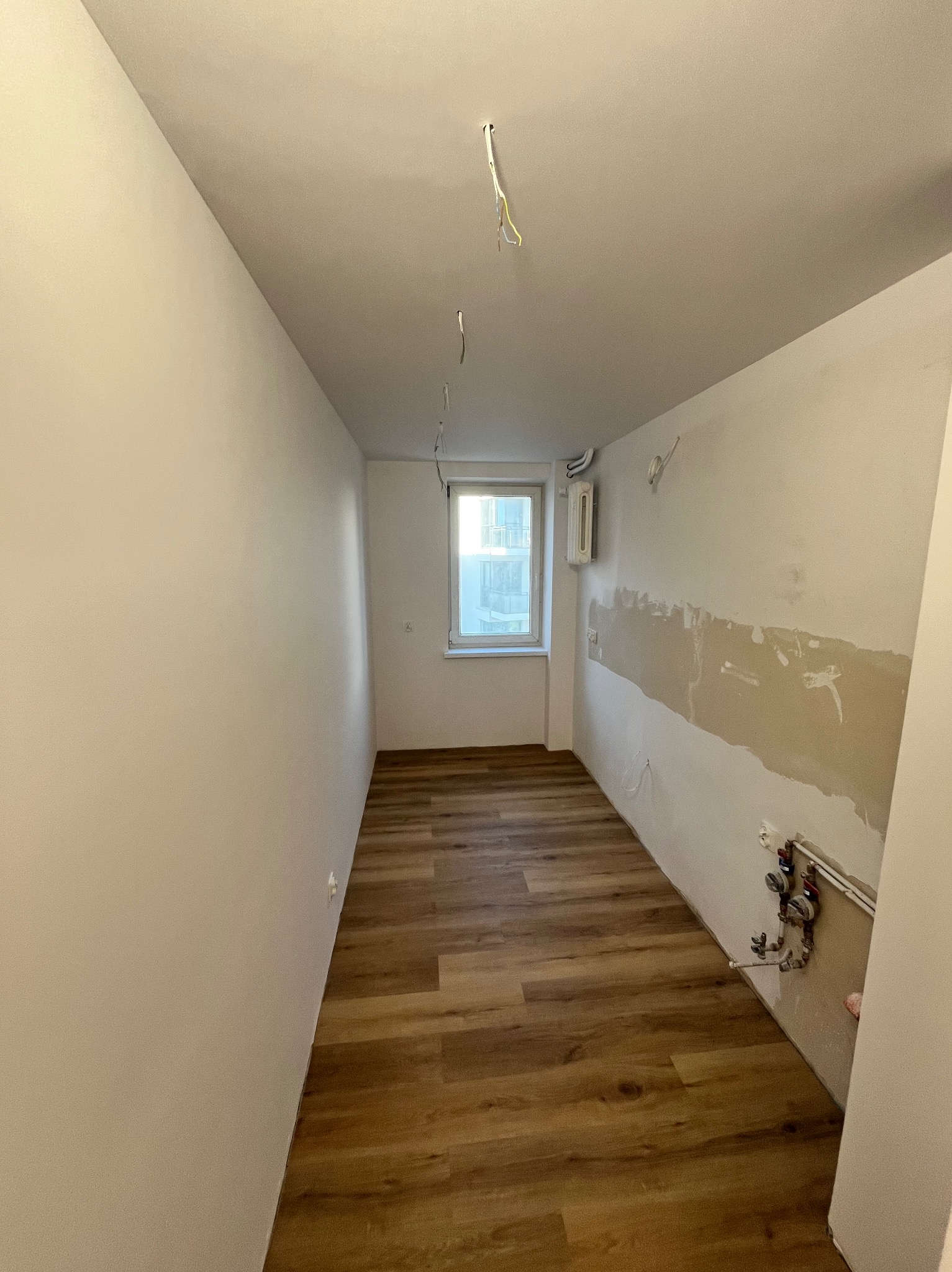 Wnętrze pokoju po remoncie: świeżo położona podłoga, częściowo wyszpachlowana ściana z instalacją wodną, wystające przewody elektryczne z sufitu, okno z widokiem.