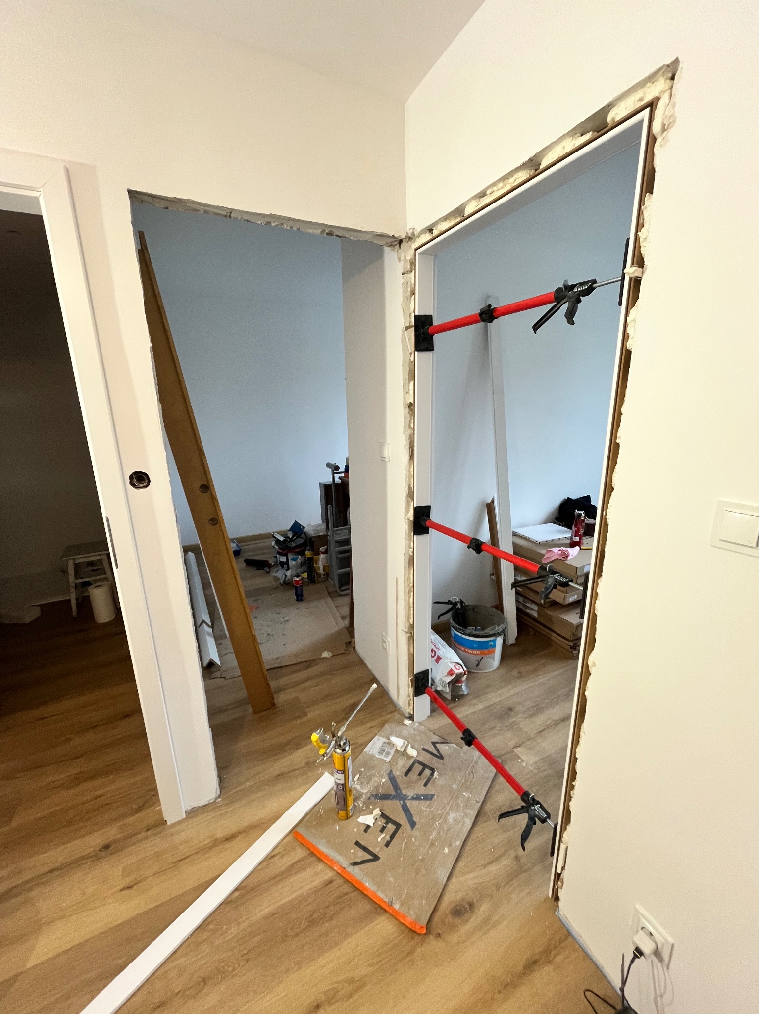 Montaż ościeżnicy drzwiowej z użyciem rozpórek teleskopowych. Widoczna piana montażowa, narzędzia i elementy wykończeniowe na podłodze. Ujęcie z perspektywy podłogi.