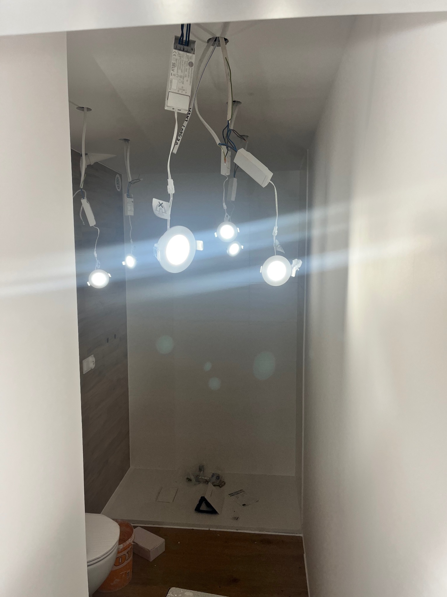 Nowoczesne oświetlenie LED w łazience w trakcie montażu, widoczne przewody i zasilacze. Jasne światło punktowe. Częściowo widoczna toaleta i surowe wykończenie.