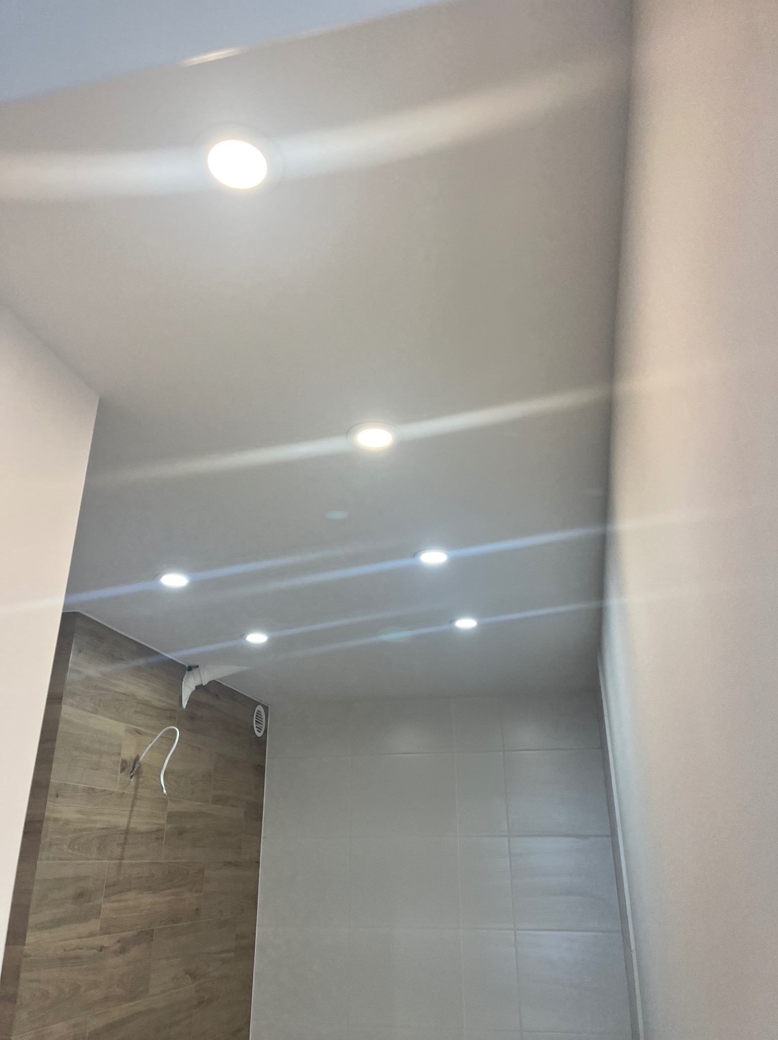 Nowoczesna łazienka z oświetleniem sufitowym LED, drewnopodobnymi płytkami i jasnoszarymi ścianami. Widoczne detale instalacji elektrycznej.