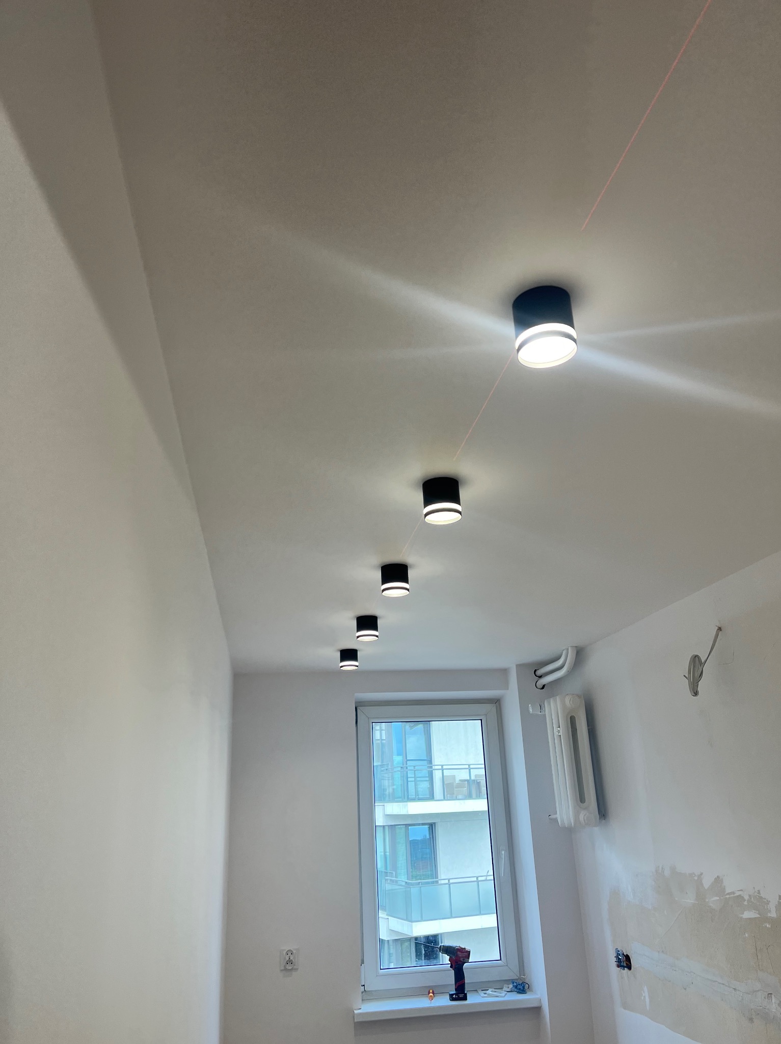 Równomiernie rozmieszczone, okrągłe lampy sufitowe w nowym pomieszczeniu, wyznaczony poziom laserem. Widoczne ślady po szpachlowaniu ścian i okno z widokiem na budynek.