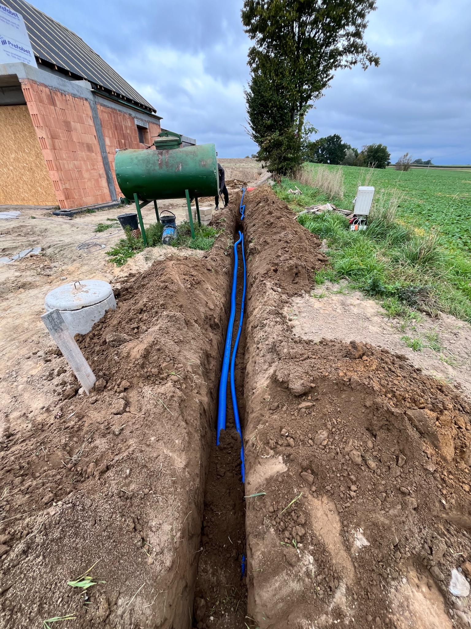 Wykop z ułożonymi, niebieskimi rurami na działce budowlanej z widoczną zielenią. W tle zbiornik i budynek w trakcie budowy pod pochmurnym niebem.