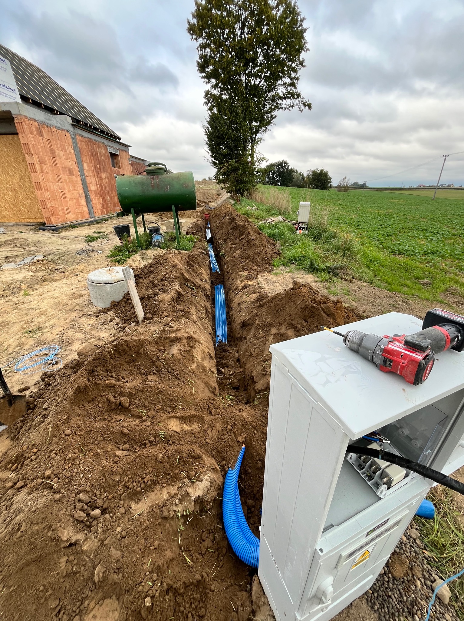 Wykop z niebieskimi rurami kablowymi w ziemi, obok biała skrzynka z narzędziami, w tle zielone pole i budynek w trakcie budowy. Instalacja podziemna.