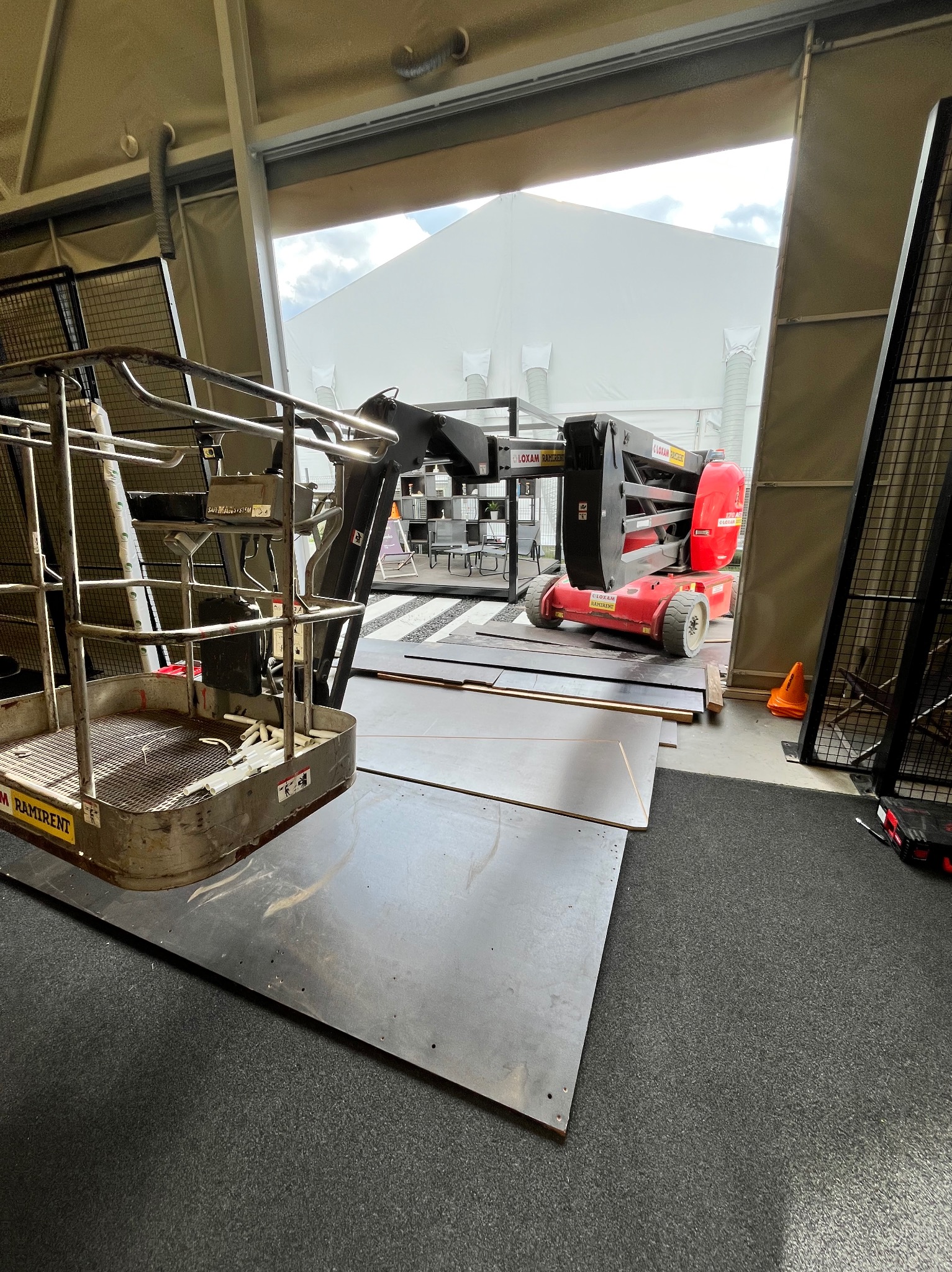 Podnośnik koszowy marki Loxam Ramirent w hali z widocznym białym namiotem w tle. Na podłodze leżą płyty. Pomarańczowy pachołek.