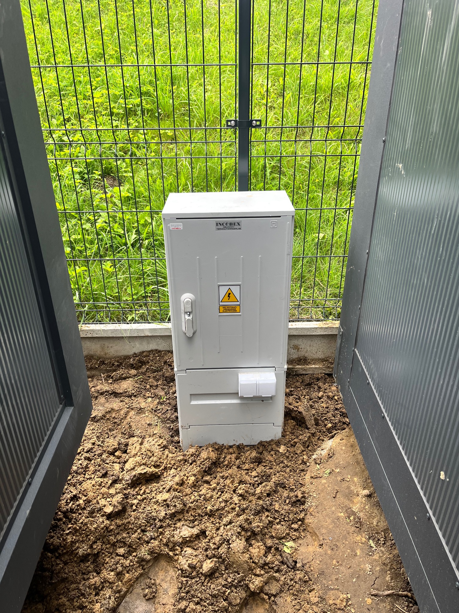 Zamontowana, biała skrzynka elektryczna IncoBex z symbolem ostrzegawczym, umieszczona na ziemi przy ogrodzeniu i otwartych, szarych drzwiach.