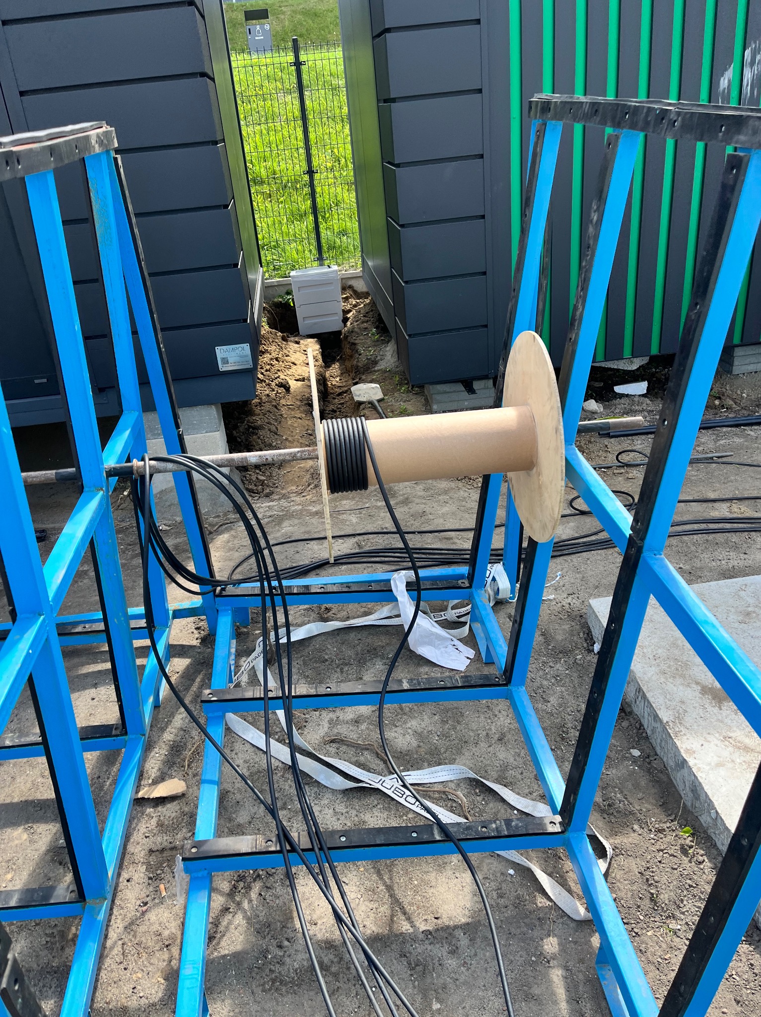 Instalacja kabli w ziemi z użyciem bębna na ramie. Widoczne czarne kable, niebieska rama, ziemia i fragment budynku w tle. Ujęcie z bliska, w dziennym oświetleniu.