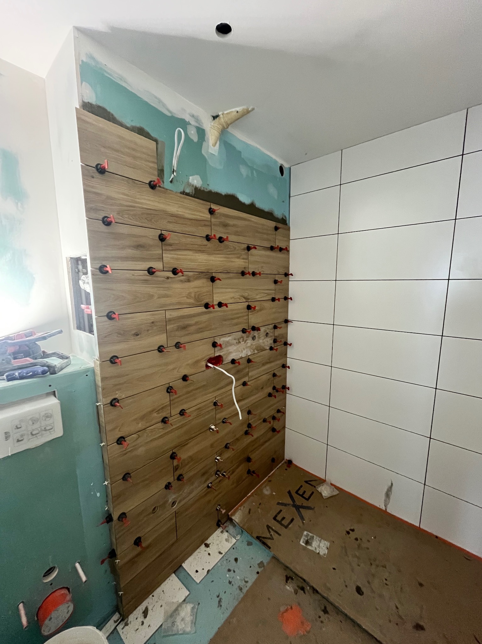 Układanie drewnopodobnych płytek z użyciem klipsów poziomujących, obok ściana z białych płytek. Widoczne narzędzia i niedokończone prace wykończeniowe.