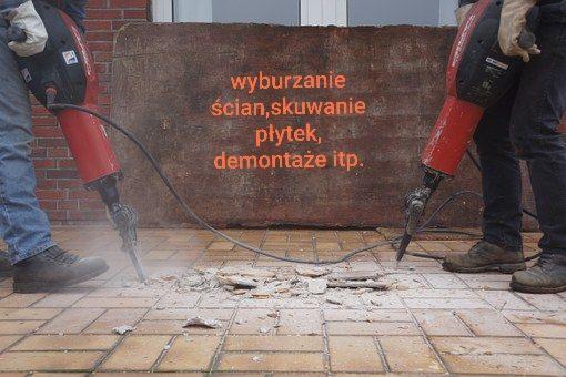 Dwóch pracowników używa młotów pneumatycznych do rozbijania brązowych płytek podłogowych na zewnątrz budynku z cegły.