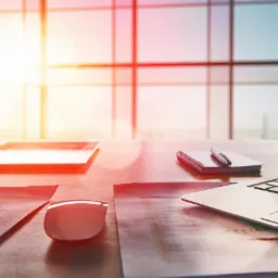 Biuro w Cieszynie w promieniach słońca: laptop, tablet, notatnik z długopisem i dokumenty na biurku przed oknem.