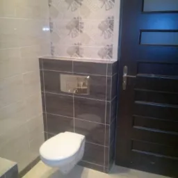 Nowoczesna toaleta podwieszana w łazience z szaro-beżowymi płytkami i ciemnymi drzwiami z przeszkleniami.