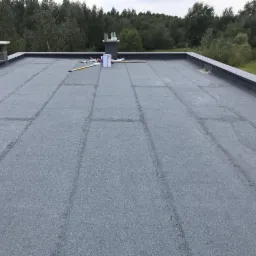 Płaski dach pokryty papą termozgrzewalną, widoczne łączenia między pasami papy, komin i narzędzia pracy w tle, drzewa w oddali.