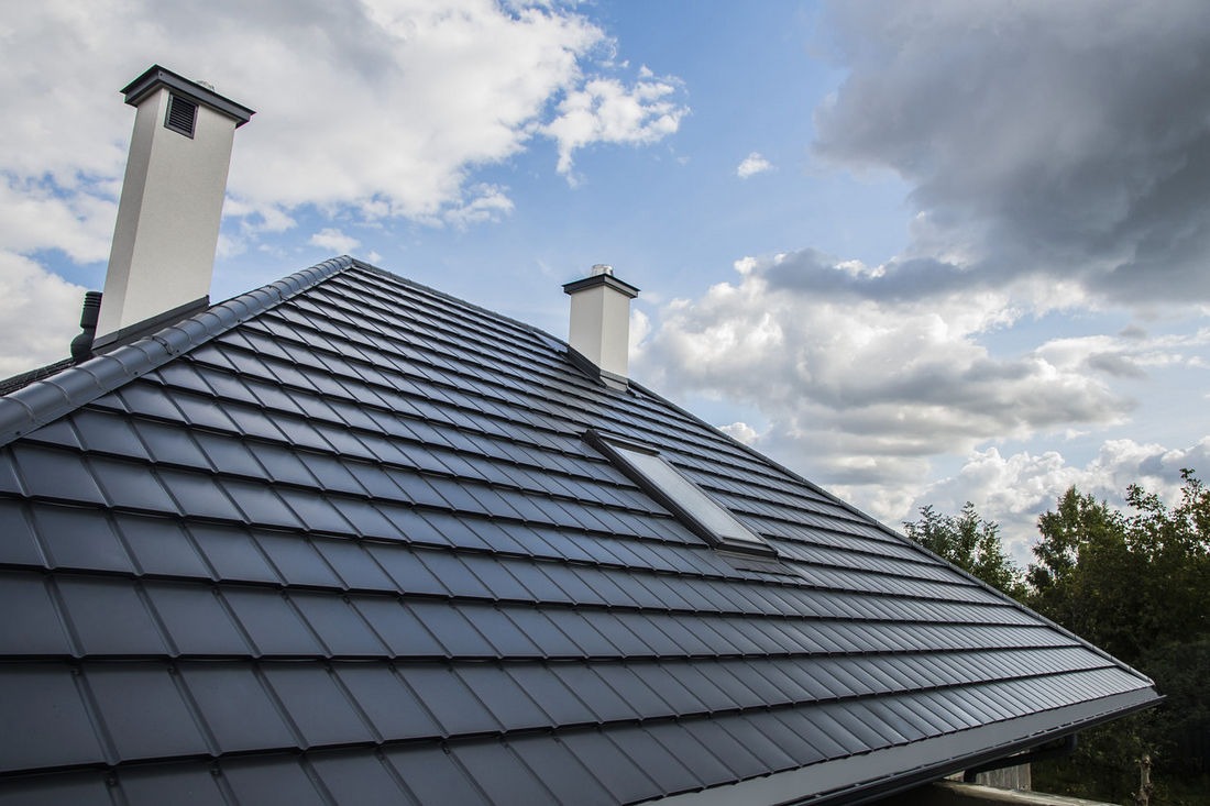 Nowoczesny, grafitowy dach z dachówki ceramicznej z oknem dachowym i dwoma kominami na tle błękitnego nieba z chmurami.