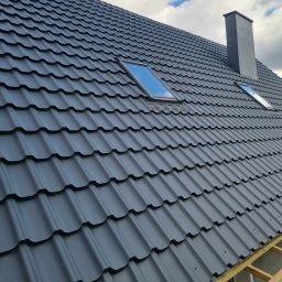 Nowoczesny, grafitowy dach z blachodachówki z dwoma oknami dachowymi i kominem w kolorze dachu, widoczne fragmenty konstrukcji drewnianej, pochmurne niebo w tle.