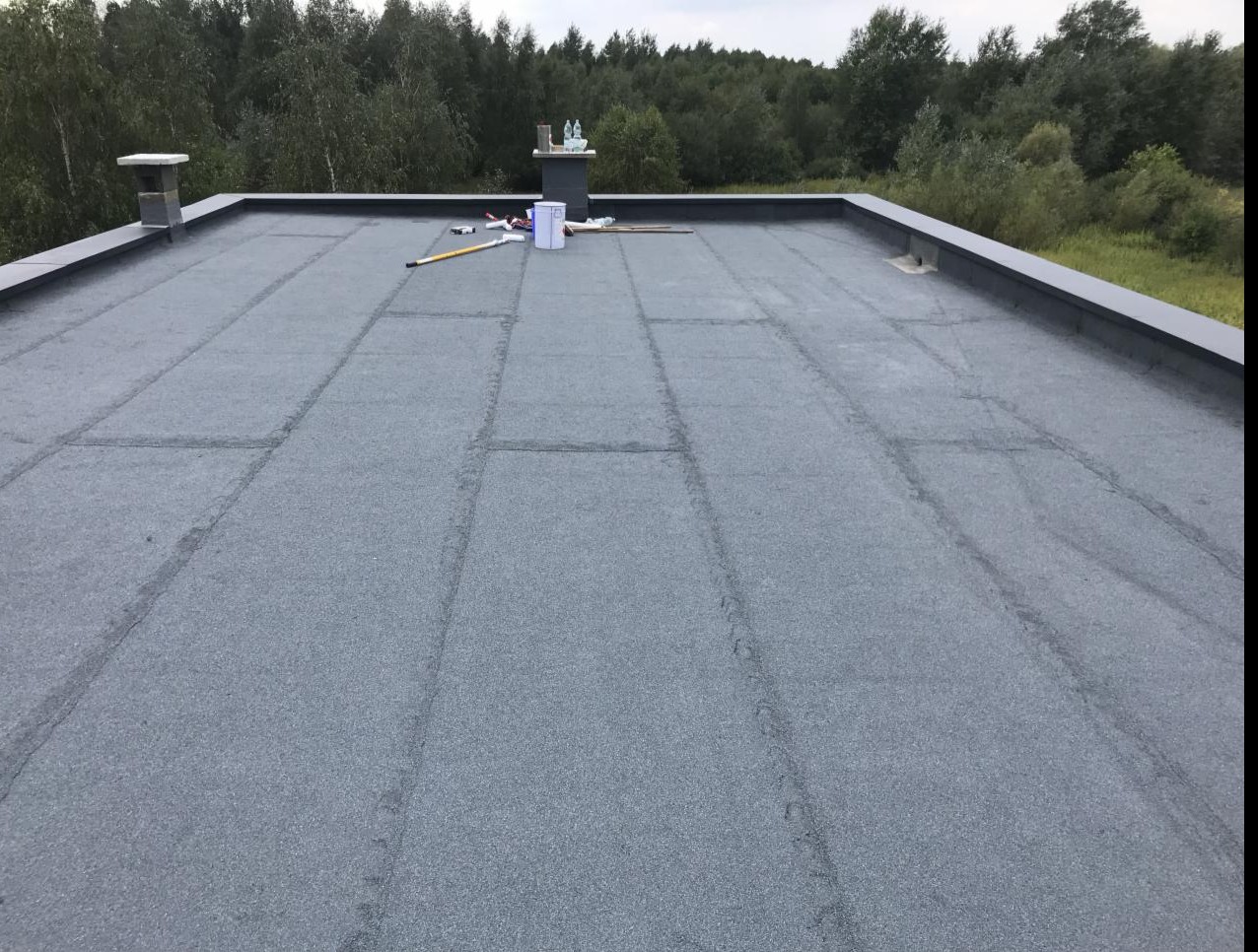 Płaski dach pokryty papą termozgrzewalną, widoczne łączenia między pasami papy, komin i narzędzia pracy w tle, drzewa w oddali.