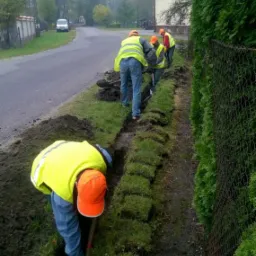 Grupa pracowników w odblaskowych kamizelkach i pomarańczowych kaskach kopie rów wzdłuż drogi i ogrodzenia, przygotowując teren pod instalację. Widoczne wykopane fragmenty darni ułożone obok rowu.