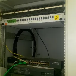 Szafa telekomunikacyjna z patch panelem, switchem Ethernet i uporządkowanymi kablami, widoczne oznaczenia portów i przewody światłowodowe.