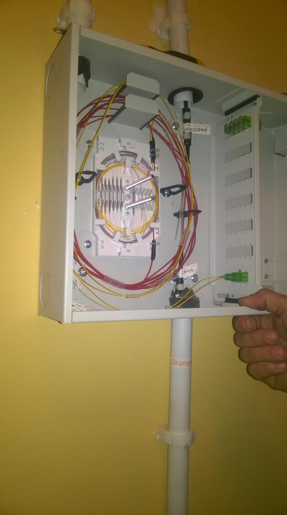 Otwarta skrzynka telekomunikacyjna z kablem światłowodowym wewnątrz, widoczna ręka przytrzymująca dolną klapę, rura oznaczona napisem 'Orange'.