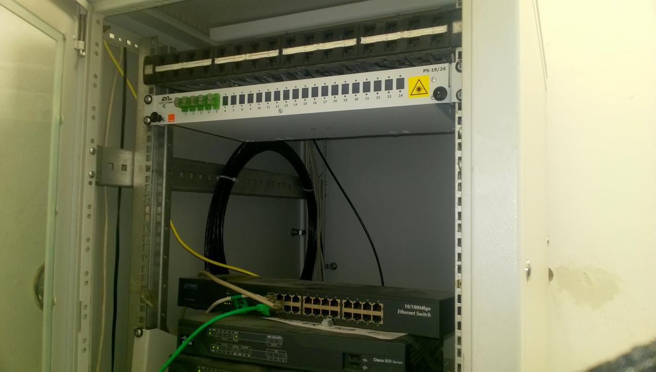 Szafa telekomunikacyjna z patch panelem, switchem Ethernet i uporządkowanymi kablami, widoczne oznaczenia portów i przewody światłowodowe.