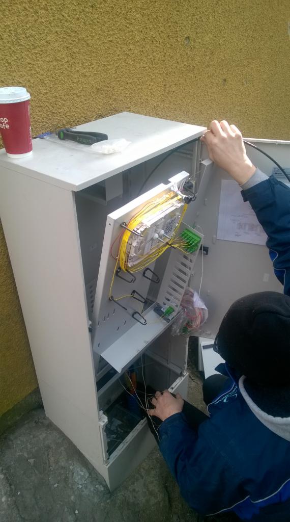 Otwarta szafka telekomunikacyjna z widocznymi światłowodami i elementami złącznymi, obok kubek po kawie i narzędzia na górnej powierzchni szafki, instalator w trakcie pracy przy podłączeniach.