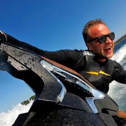 Fabryka Adrenaliny - Mężczyzna w okularach przeciwsłonecznych, w czarnej kamizelce z żółtym paskiem, z szerokim uśmiechem pędzi na skuterze wodnym po błękitnej wodzie, z widokiem na zielony brzeg w oddali.