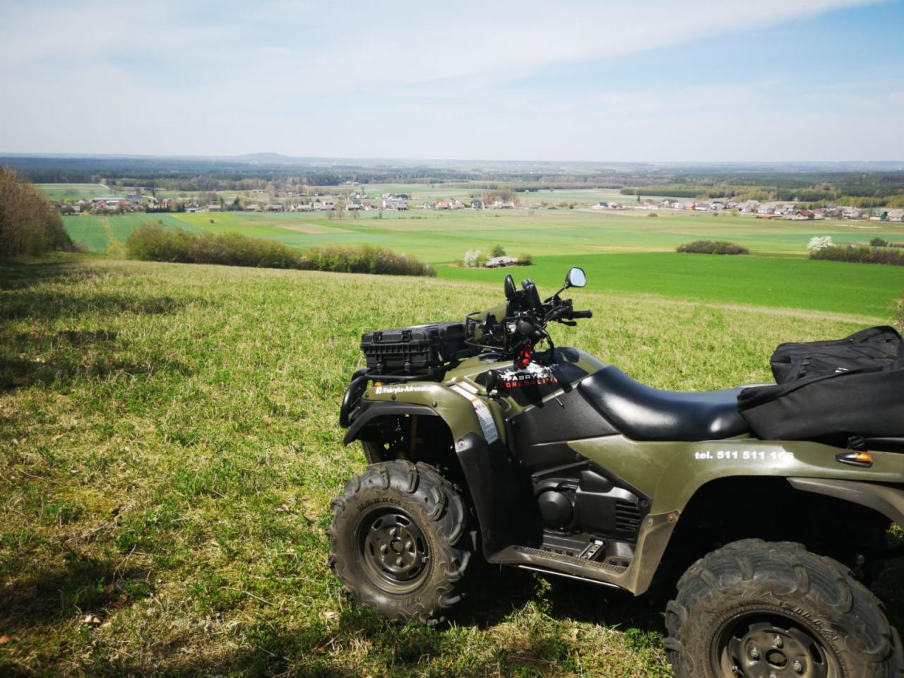 Zielony quad ATV zaparkowany na wzgórzu z widokiem na wiejski krajobraz z polami i zabudowaniami w tle, błękitne niebo.
