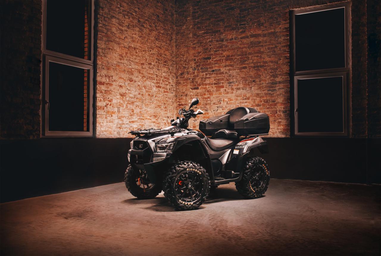 Czarny quad Kymco z oponami Maxxis Bighorn, zaparkowany w narożniku pomieszczenia z ceglaną ścianą i oknami.