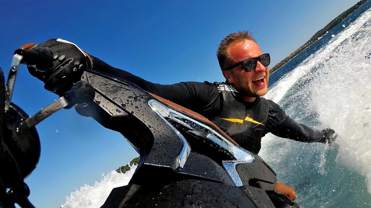 Mężczyzna w okularach przeciwsłonecznych, w czarnej kamizelce z żółtym paskiem, z szerokim uśmiechem pędzi na skuterze wodnym po błękitnej wodzie, z widokiem na zielony brzeg w oddali.