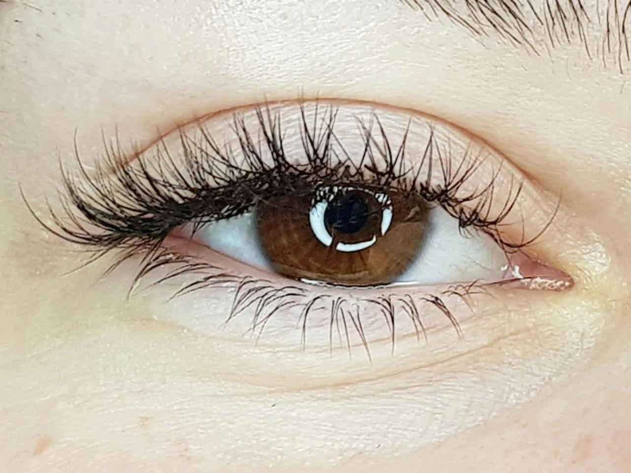 Zbliżenie na oko z brązową tęczówką i przedłużonymi, czarnymi rzęsami, widoczne delikatne odbicie światła w źrenicy.