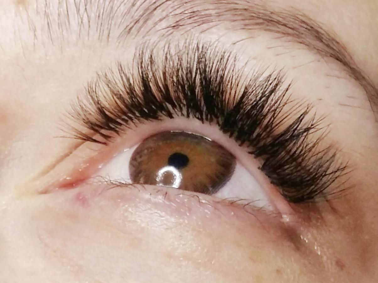 Zbliżenie na oko z brązową tęczówką i gęstymi, czarnymi, profesjonalnie przedłużonymi rzęsami. Widoczny delikatny rumień na powiece.