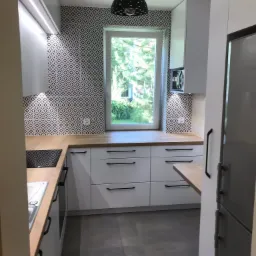 Nowoczesna kuchnia z białymi szafkami z czarnymi uchwytami, drewnianym blatem, geometryczną tapetą i widokiem na zieleń za oknem.