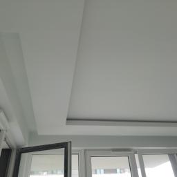 6th Avenue - Nowoczesny sufit podwieszany z płyt kartonowo-gipsowych, białe ściany i częściowo otwarte okno balkonowe z widokiem na miasto.