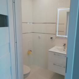 6th Avenue - Nowoczesna, biała łazienka z podświetlanym lustrem, umywalką z szafką i toaletą, widoczna przez częściowo otwarte drzwi.