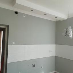 6th Avenue - Szare ściany z białymi płytkami dekoracyjnymi do połowy wysokości, widoczne gniazdka elektryczne, włącznik światła, otwór w suficie, lampy wiszące jeszcze w folii ochronnej i częściowo otwarte drzwi.