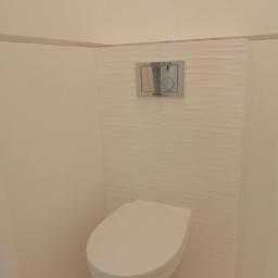 6th Avenue - Podwieszana toaleta z białym siedziskiem i chromowanym przyciskiem spłukującym na ścianie z dekoracyjnymi, falistymi płytkami w minimalistycznej, jasnej łazience.