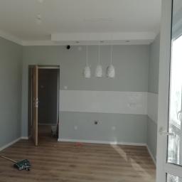 6th Avenue - Pokój po odświeżeniu: szare ściany z białym pasem dekoracyjnym, nowe lampy wiszące w folii ochronnej, drewnopodobna podłoga, otwarte drzwi i narzędzia malarskie.